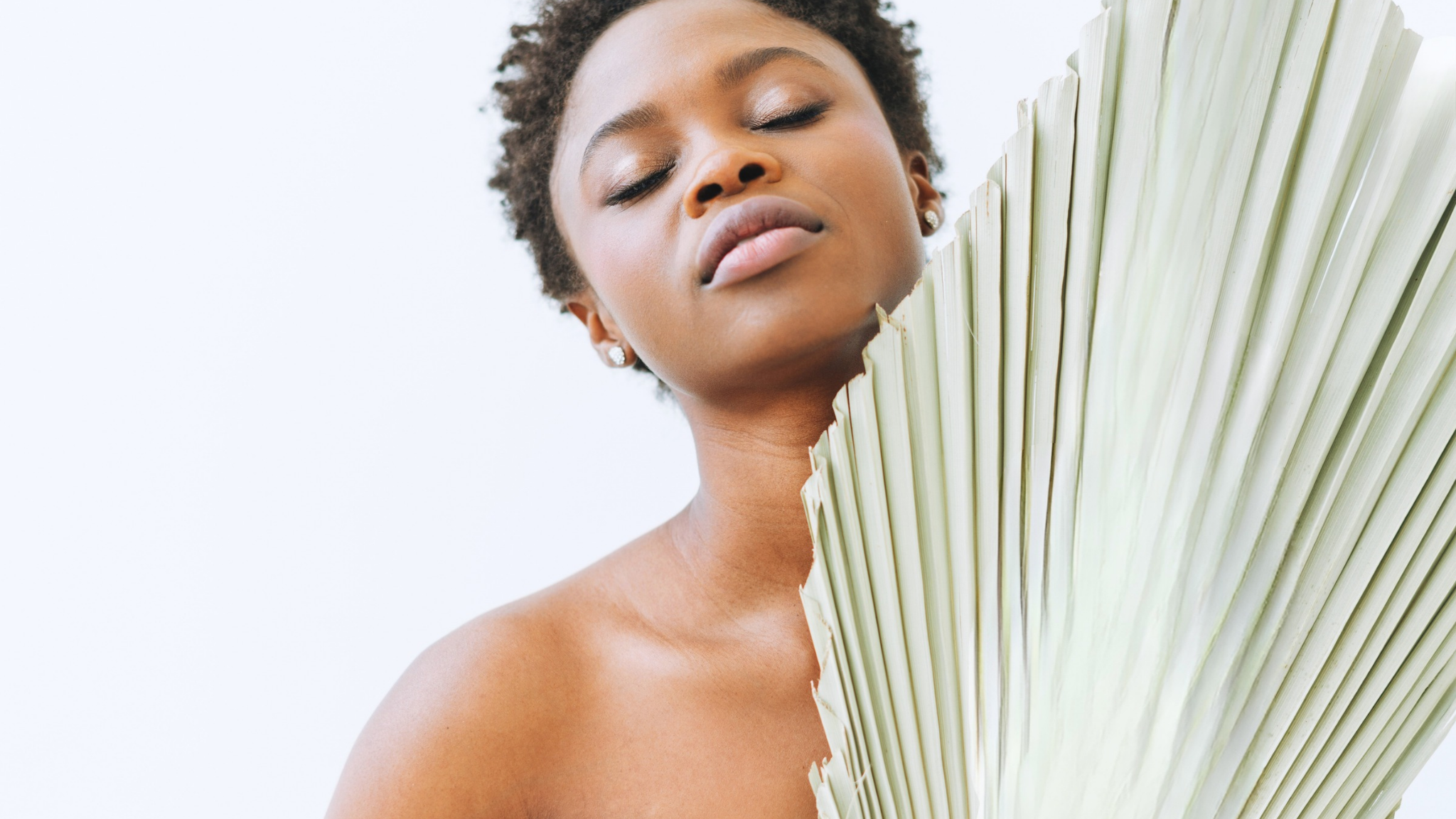 A close-up lifestyle shot of a woman with short curly hair, her eyes closed, holding a large, light sage green palm fan next to her face against a clean white background.