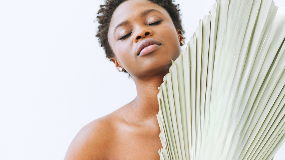 A close-up lifestyle shot of a woman with short curly hair, her eyes closed, holding a large, light sage green palm fan next to her face against a clean white background.